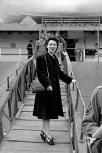 A young white woman in a long black coat stands on a ramp to a boat.