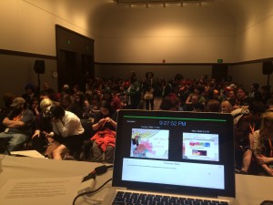 A shot from behind the presenter's computer, full view of the crowd in a room.