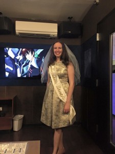 A young woman in a silver dress and white veil stands in front of a TV with an image from Gundam Seed