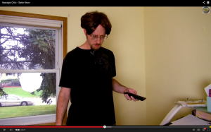 Nostalgia Critic stands holding a remote wearing a black t-shirt and pimples on his face.