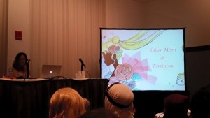 A young white woman sits at a table with a mic with a big screen next to her with the words Sailor Moon & Feminism