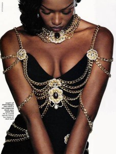 Naomi Campbell models a black dress with gold chains draped around the arms and chest