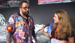 A bearded white man wearing a shirt with very sexy white women on it stands next to a young woman with a microphone