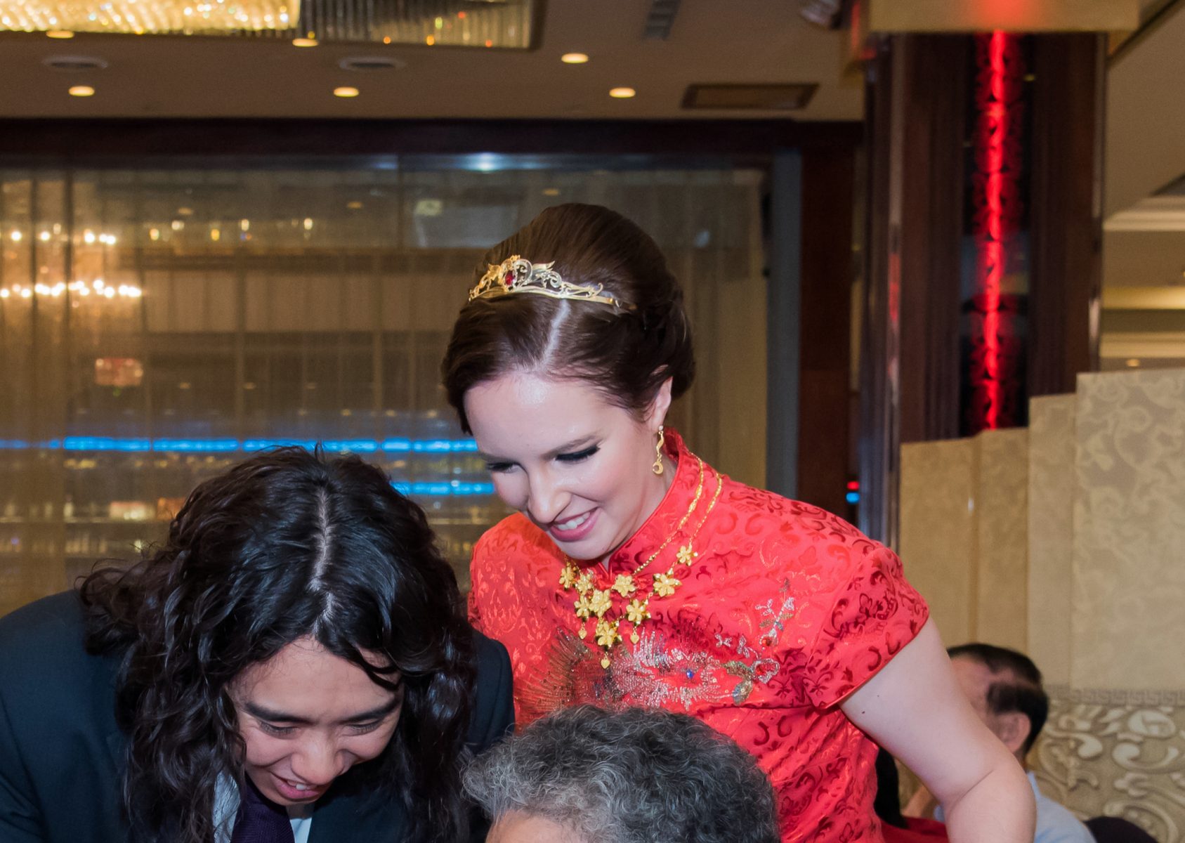 a young white woman wears a red qipao with a gold flower necklace, gold earrings and a gold tiara with a red stone